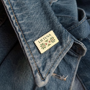 May include: A white and gold pin with a green and red design. The pin features the word "LIETUVA" and a traditional Lithuanian pattern.