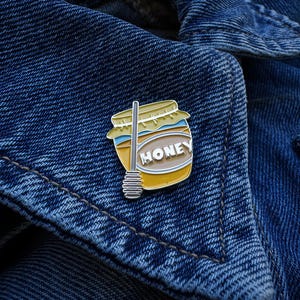 May include: An enamel pin depicting a honey jar with a silver-coloured honey dipper. The pin features a yellow jar with a blue and white label that reads "HONEY". The pin is set against a blue denim jacket.