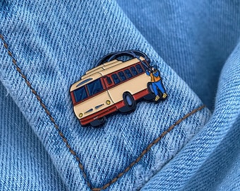 May include: A white and red enamel pin of a trolley bus with a person holding onto the overhead wire. The pin is on a blue denim jacket.