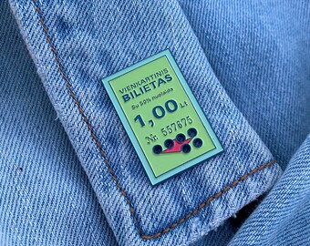 May include: A blue and green enamel pin shaped like a ticket with the text "Vienkartinis Bilietas" and "Su 50% nuolaida" and the number "1,00 Lt" and "Nr. 557675".