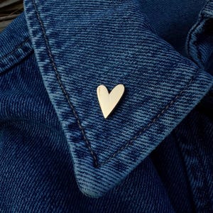 Könnte beinhalten: Eine polierte, goldfarbene herzförmige Anstecknadel am Kragen einer dunkelblauen Jeansjacke. Das Herz hat eine glatte, reflektierende Oberfläche und eine spitze Unterseite, die einen Kontrast zum strukturierten Stoff der Jeansjacke bildet.