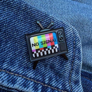 Könnte beinhalten: Ein schwarzer Emaille-Pin in Form eines Retro-Fernsehers. Der Bildschirm zeigt ein farbenfrohes Testmuster mit den Worten "NO SIGNAL". Der Pin hat zwei Antennen und ist vor einem blauen Jeanshintergrund platziert.