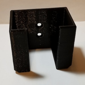 May include: Black plastic wall mount with two holes for mounting screws. The mount has a rectangular opening for holding an object.
