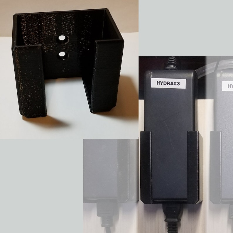 May include: A black 3D-printed wall mount for an electronic device. The mount has a rectangular shape with two holes and two side supports. The text "HYDRA#3" is visible on the device.