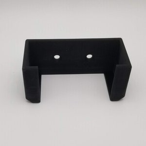 May include: Black plastic wall mount with two holes for mounting screws. The mount has a U-shaped opening for holding an object.
