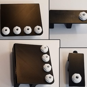May include: A black plastic 3D printed wall mount with five white plastic knobs. The mount has a rectangular shape with a slight angle on the top edge. The knobs are evenly spaced along the bottom edge of the mount.