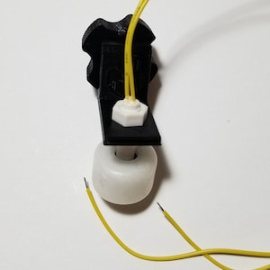 May include: A white float switch with two yellow wires attached to a black plastic mounting bracket. The float switch is designed to detect the level of liquid in a tank or container.