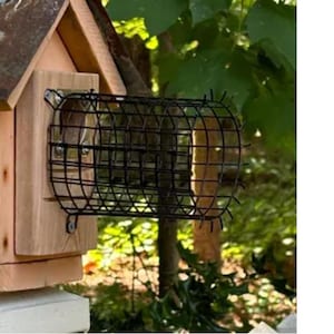 May include: A wooden bird feeder with a metal roof and a black wire cage. The birdhouse is made of light brown wood and is mounted on a white stone base. The background features green foliage.