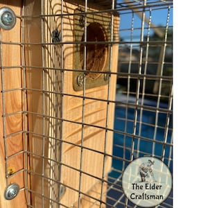 May include: Close-up of a wooden birdhouse with a wire mesh front. The birdhouse is made of light brown wood and has a circular entrance. The wire mesh is attached with screws. The Elder Craftsman logo is in the bottom right corner.