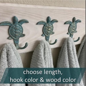 May include: Three teal turtle-shaped hooks mounted on a light wood board, each holding a gray towel. The hooks have a distressed finish. The text at the bottom says "choose length, hook color & wood color."