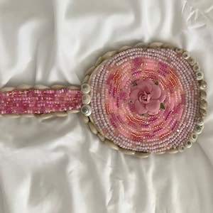 Puede incluir: Un espejo de mano de cuentas rosa y blanco con una flor rosa en el centro. El espejo está rodeado por un borde de conchas blancas.