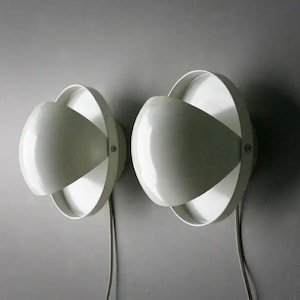 May include: A pair of white wall-mounted lamps featuring a circular outer ring and a smaller, rounded inner dome shade. Each lamp is secured with a visible metal screw on the side and includes a white power cord extending from the bottom base.