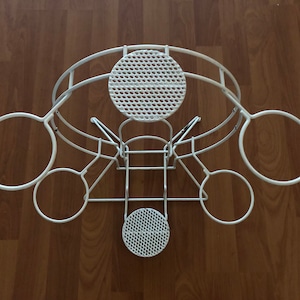 May include: White metal plant holder with a unique design. It features circular and semi-circular holders, along with two perforated circular platforms. The structure is made of thin, curved metal rods, creating an open and airy aesthetic.