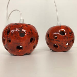 Könnte beinhalten: Zwei runde, rote Keramik-Pendelleuchten mit mehreren kreisförmigen Öffnungen. Die Leuchten haben eine strukturierte Oberfläche und werden von weißen Kabeln abgehängt. Die Leuchten sind leuchtend rot mit dunkelbraunen Akzenten.