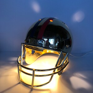 May include: A gold football helmet lamp with a red, white, and blue stripe design. The helmet has a clear light bulb inside and a metal face mask.