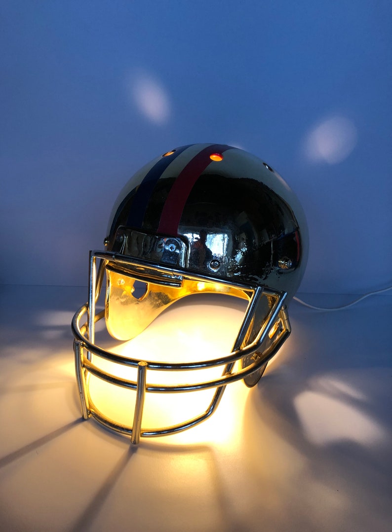 May include: A gold football helmet lamp with a red, white, and blue stripe design. The helmet has a clear light bulb inside and a metal face mask.