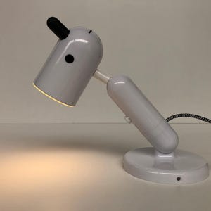 May include: A white desk lamp with a black accent on the top and a black and white striped cord. The lamp has a round base and an adjustable arm. The lamp is lit, casting a warm glow.