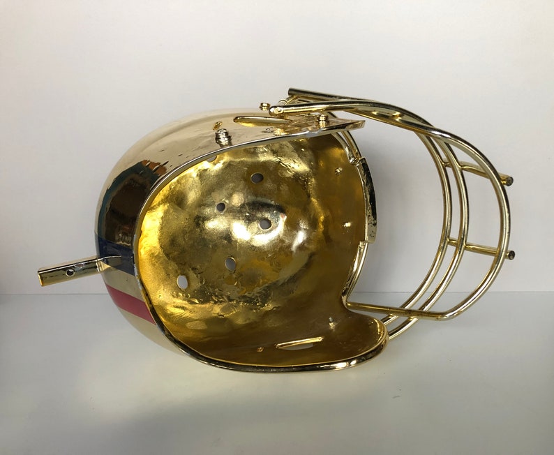 May include: A gold-colored football helmet with a red, white, and blue stripe design. The helmet is open on the top and has a wire face mask.