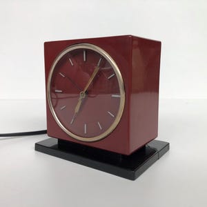 May include: A square, deep red electric desk clock with a circular face, thin gold-tone bezel, and simple white hour markers. The clock is mounted on a rectangular black base and includes a black power cord extending from the side of the unit.