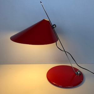 May include: A red desk lamp with a conical shade and a chrome arm. The lamp has a round base and a black cord.