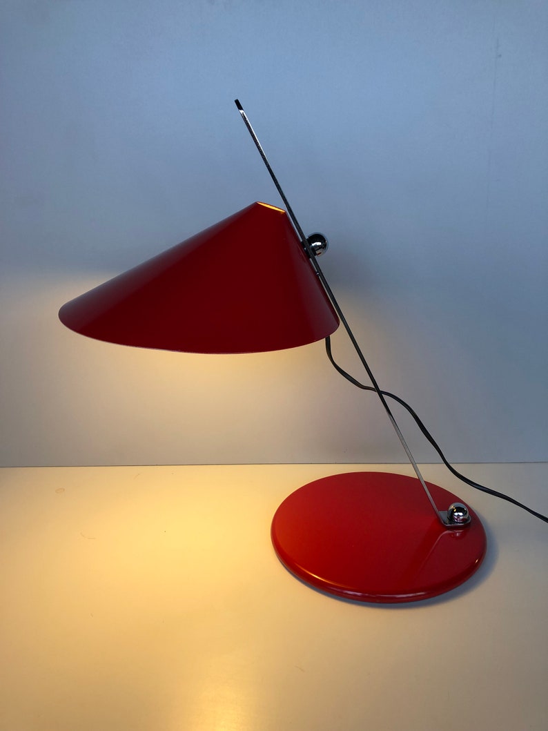 May include: A red desk lamp with a conical shade and a chrome arm. The lamp has a round base and a black cord.
