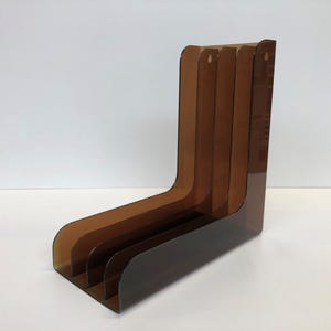 以下が含まれることがあります： 4つの区画を持つ茶色のアクリル製雑誌ホルダー。ホルダーはL字型で、モダンなデザインです。