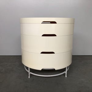 May include: A white, round, three-drawer nightstand with brown handles. The nightstand is on a white metal base.