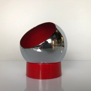 May include: A chrome and red bowl with a half-sphere shape. The bowl is tilted at an angle and sits on a red base.