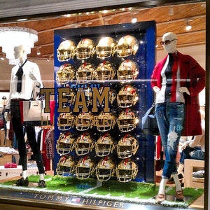 May include: A storefront display featuring a mannequin wearing a red and white striped shirt and blue jeans. The mannequin is standing in front of a wall of gold football helmets with the word "TEAM" in large gold letters. The storefront is decorated with green grass and a sign that reads "Tommy Hilfiger".