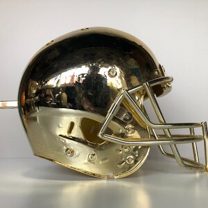 May include: A gold football helmet with a face mask. The helmet is shiny and has a smooth surface.