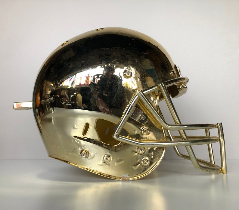 May include: A gold football helmet with a face mask. The helmet is shiny and has a smooth surface.