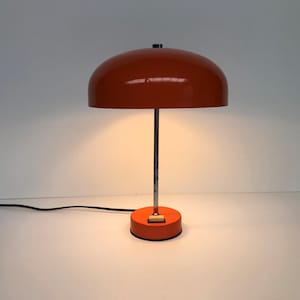 May include: A mid-century modern orange table lamp with a domed shade. The lamp has a cylindrical metal stem and a circular base. The lamp is plugged into a wall outlet with a black cord. The lamp is a retro design.
