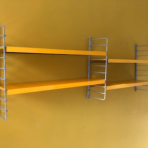 May include: A wall-mounted shelving unit with three bright yellow shelves and a silver metal frame. The shelves are rectangular and the frame has a grid-like design. The unit is mounted on a yellow wall.