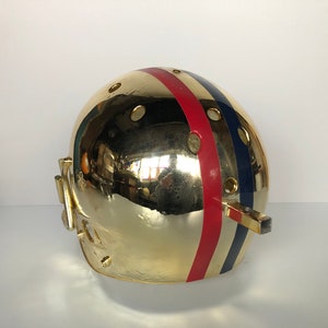 May include: A gold football helmet with red, white, and blue stripes. The helmet has a face mask and a chin strap.