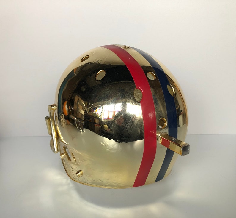 May include: A gold football helmet with red, white, and blue stripes. The helmet has a face mask and a chin strap.
