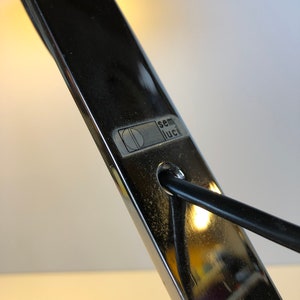 May include: Close-up of a silver desk lamp with a black cord. The lamp has a label that reads "semi luci" with a stylized letter "D" inside a circle.