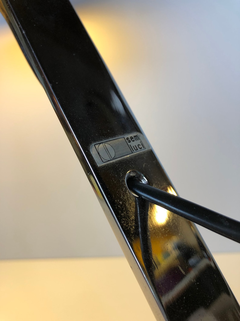 May include: Close-up of a silver desk lamp with a black cord. The lamp has a label that reads "semi luci" with a stylized letter "D" inside a circle.