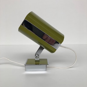 May include: Olive green and chrome desk lamp. The cylindrical lamp head is angled, with a reflective chrome band. The base is a square with a chrome frame and a green top. A white cable extends from the base.