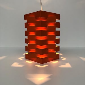 Peut inclure: Suspension luminaire géométrique orange avec un design en couches. La lumière projette des ombres triangulaires. Le luminaire est suspendu par un cordon blanc, créant un élément d'éclairage moderne et artistique.