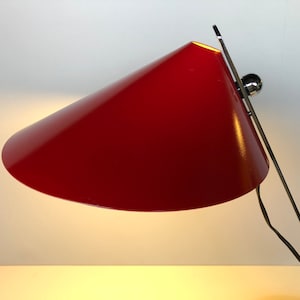 May include: A red metal desk lamp with a conical shade and a chrome base. The lamp is turned on and the light is shining.