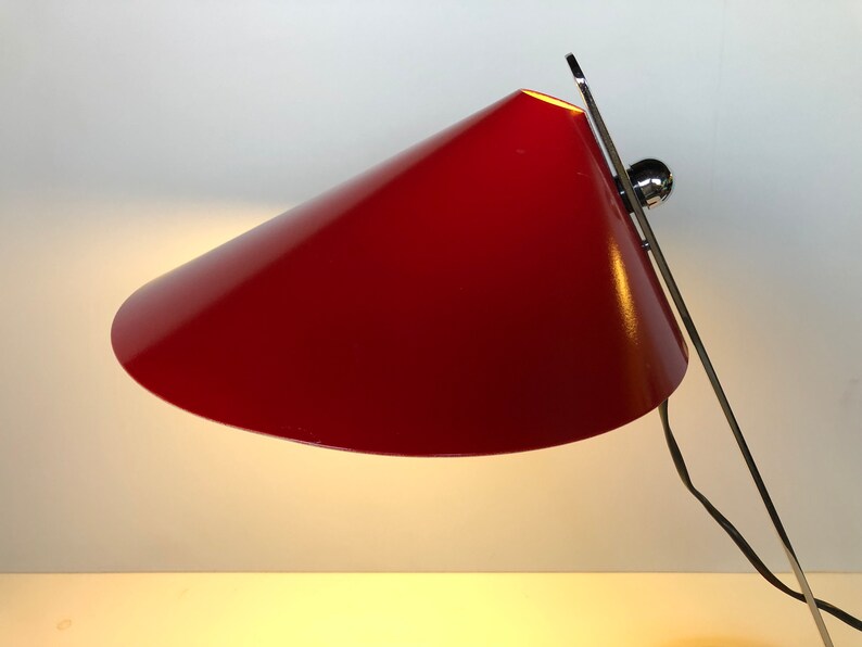 May include: A red metal desk lamp with a conical shade and a chrome base. The lamp is turned on and the light is shining.