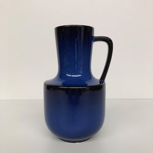 Op de afbeelding: Blauwe keramische kan met een zwarte handgreep. De kan heeft een afgeronde basis en een slanke hals. De kleur gaat over van een diepblauw aan de basis naar een donkerder blauw aan de bovenkant. De handgreep is zwart.
