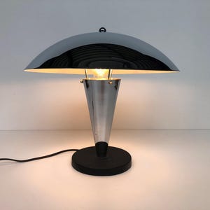 May include: A table lamp featuring a polished chrome dome-shaped shade and a conical chrome base. The lamp stands on a circular black base and emits a warm light from the center. A black power cord extends from the base of the lamp.