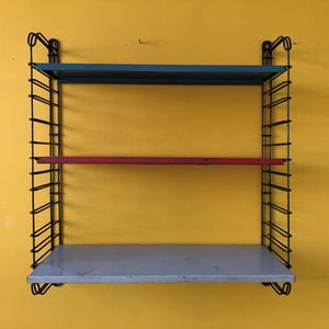 May include: A three-tiered wall-mounted shelf with a black wire frame. The shelves are painted blue, red, and gray. The shelf is mounted on a yellow wall. The shelf is a vintage design.