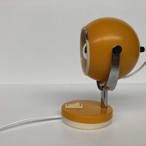 Puede incluir: Una lámpara de escritorio amarilla mostaza vintage con una cabeza esférica y una base circular. La lámpara tiene un brazo de soporte cromado y un cable de alimentación blanco. La base tiene un interruptor de encendido/apagado. La lámpara está sobre un fondo blanco.