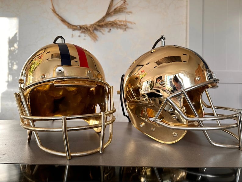 May include: Two gold football helmets with face masks. One helmet has a blue and red stripe on the side.