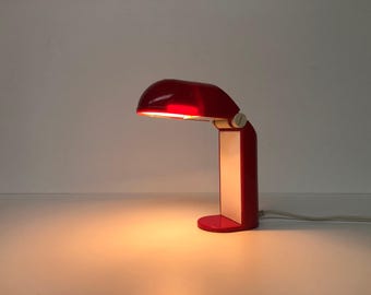 Könnte beinhalten: Eine rote Schreibtischlampe mit weißem Fuß und rotem Schirm. Die Lampe ist eingeschaltet und das Licht leuchtet.