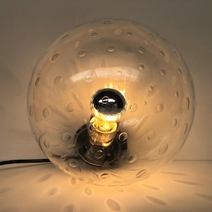 May include: A clear glass globe lamp with a decorative pattern of small circles. The lamp features a chrome-colored base and a visible light bulb. The lamp is plugged into a black cord.