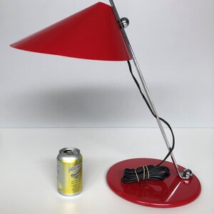 May include: A red desk lamp with a conical shade and a chrome base. The lamp is on a white surface with a can of soda in the foreground.
