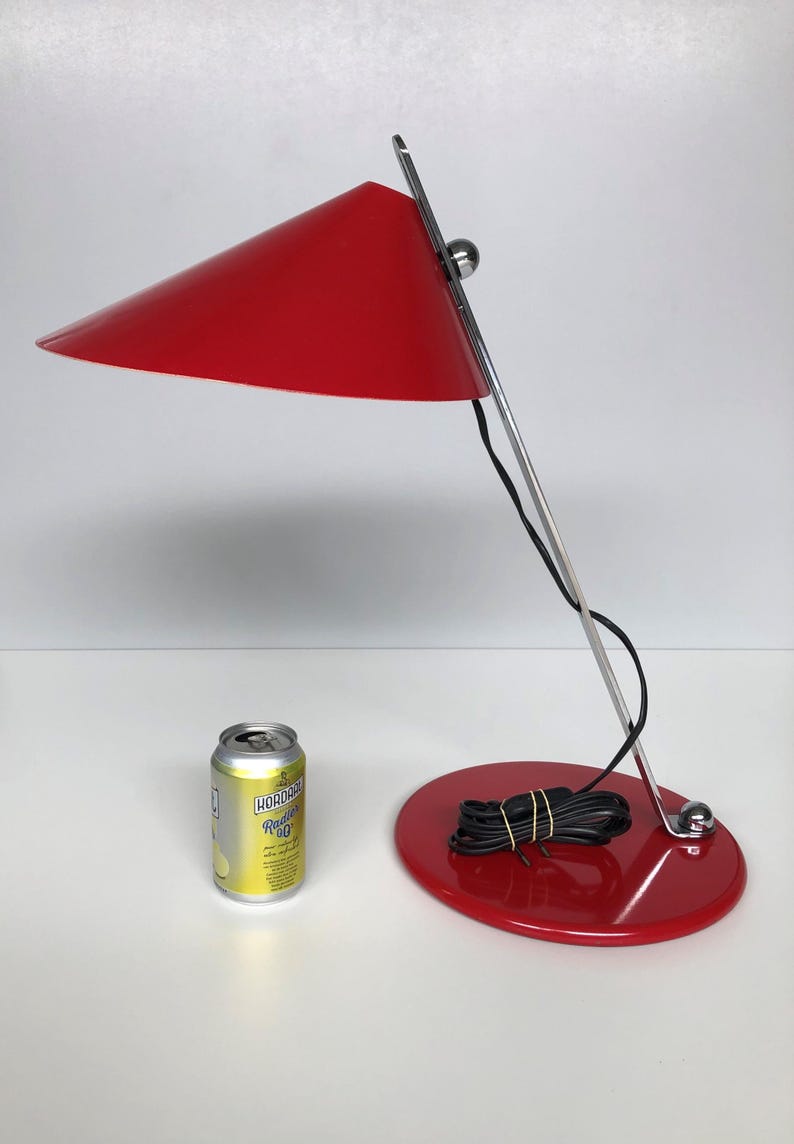 May include: A red desk lamp with a conical shade and a chrome base. The lamp is on a white surface with a can of soda in the foreground.